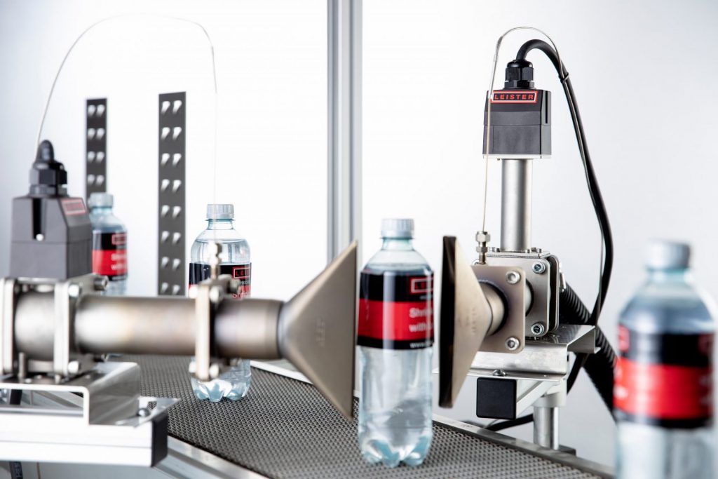 Sistema industrial con calentadores de aire Leister LHS aplicando calor preciso para el encogimiento uniforme de etiquetas en botellas durante procesos de embalaje