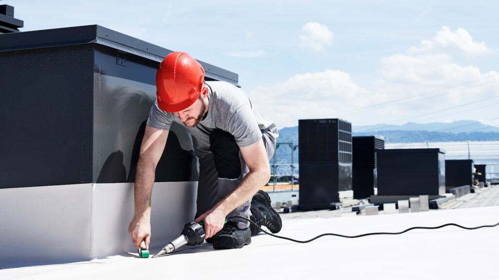 Operario en techo usando la pistola de aire caliente Leister TRIAC AT para soldar y reparar membranas plásticas en cubierta